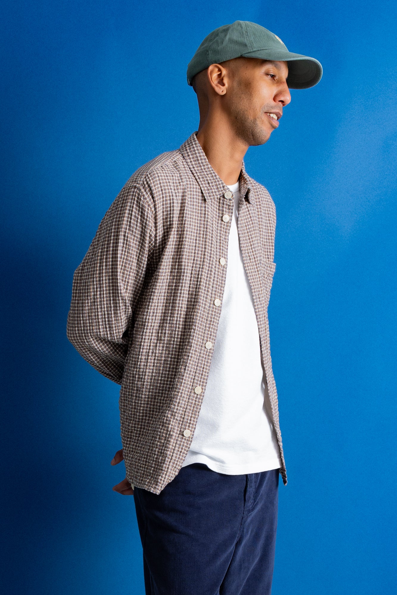 A man wearing a brown and white checkered button-down shirt over a white t-shirt, navy pants, and a green cap, standing against a solid blue background.