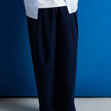 A person wearing loose dark navy trousers, a white shirt, and black shoes, standing against a solid blue wall.