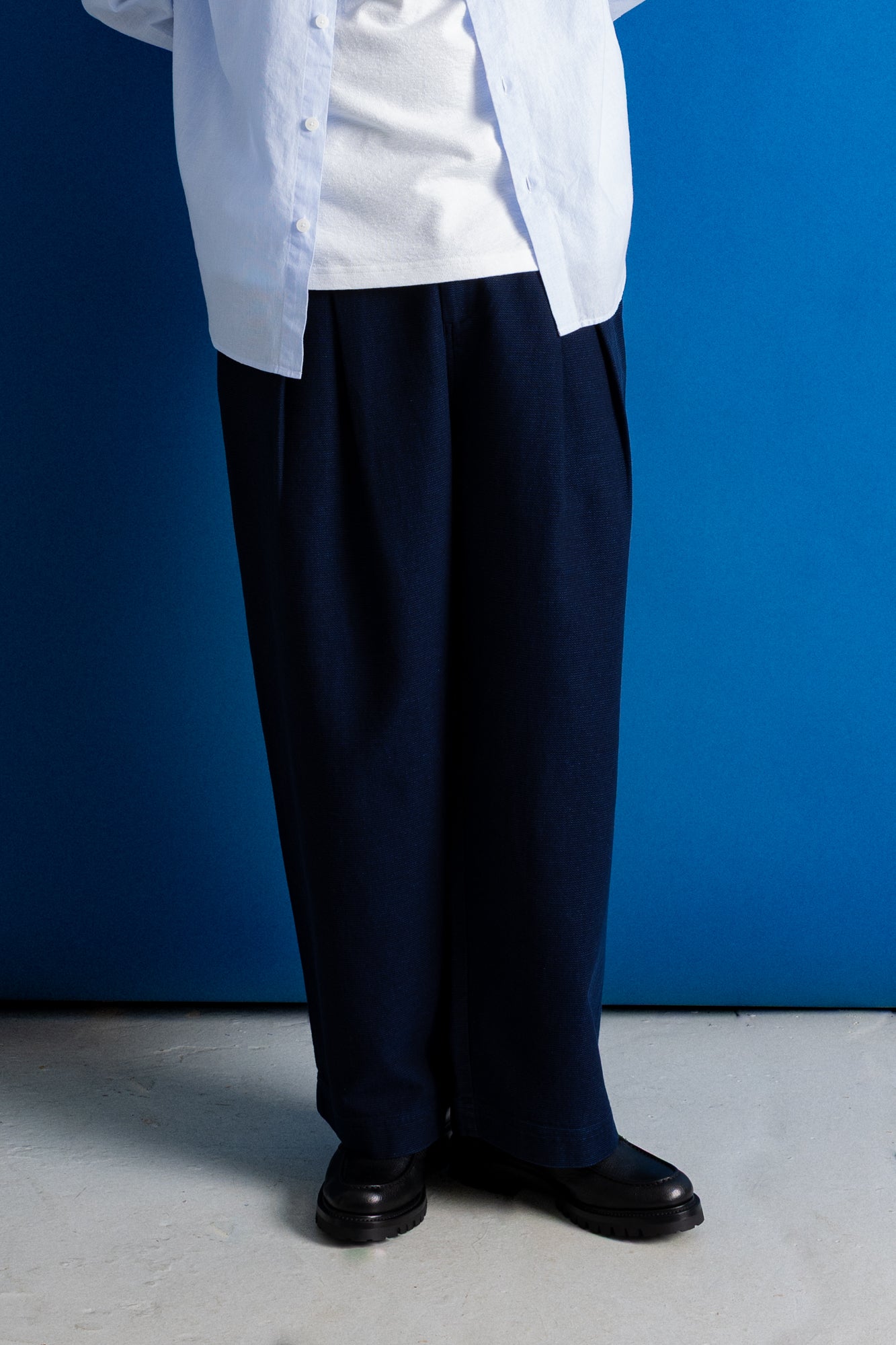 A person wearing loose dark navy trousers, a white shirt, and black shoes, standing against a solid blue wall.