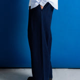 A person shown from the waist down wearing dark navy wide-leg trousers, a light blue button-up shirt, and black leather shoes, standing against a vibrant blue wall and concrete floor.
