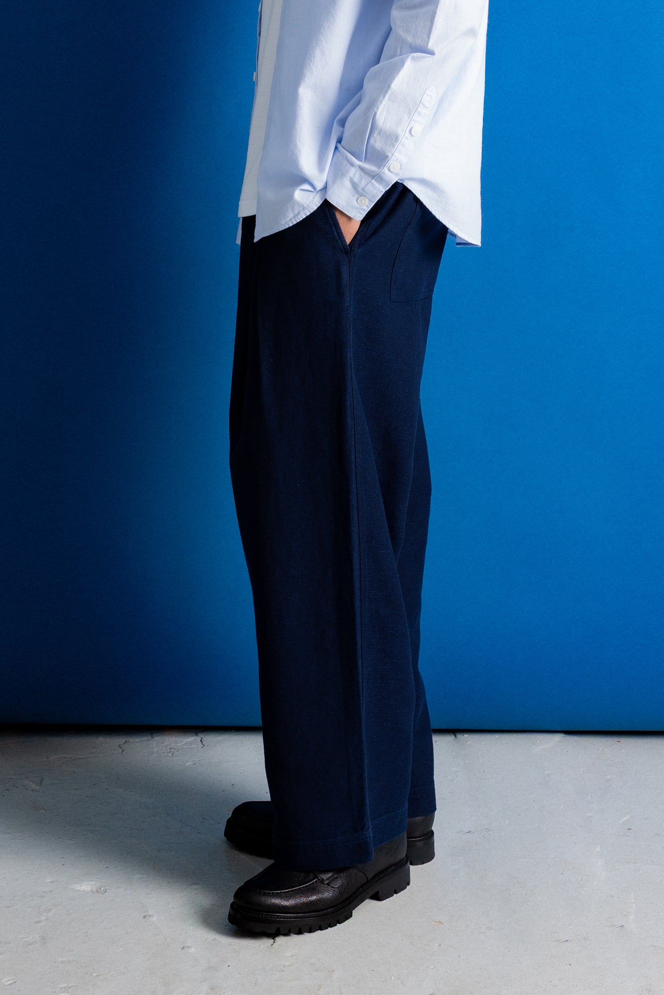 A person shown from the waist down wearing dark navy wide-leg trousers, a light blue button-up shirt, and black leather shoes, standing against a vibrant blue wall and concrete floor.