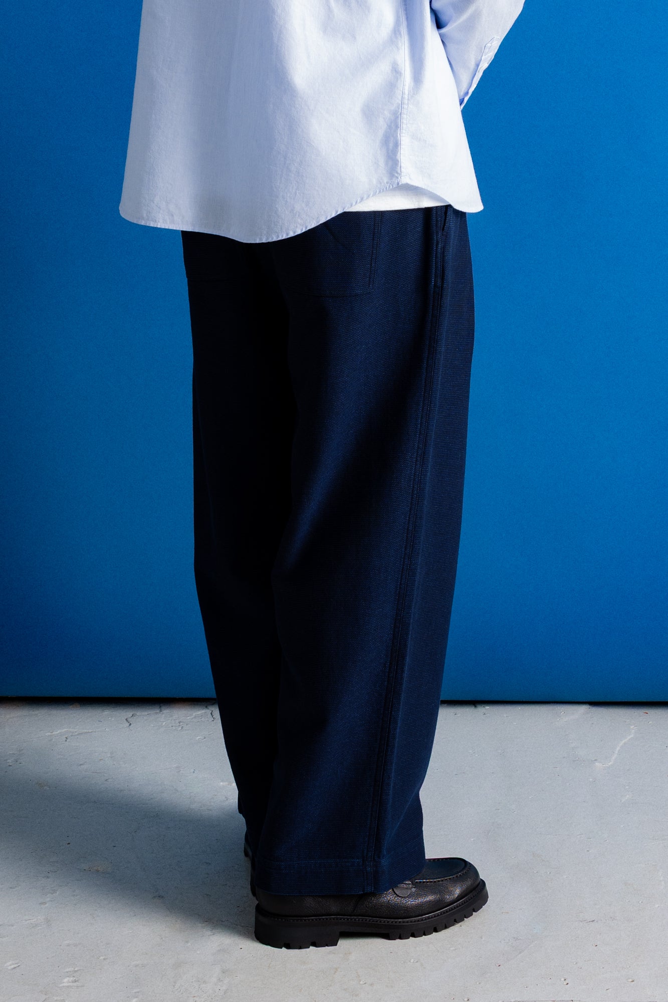 A person shown from the waist down wearing dark navy wide-leg trousers, a light blue shirt, and black shoes, standing against a solid blue wall.