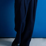 A person wearing loose dark navy pleated trousers, a white shirt, and black shoes, standing against a solid blue wall.