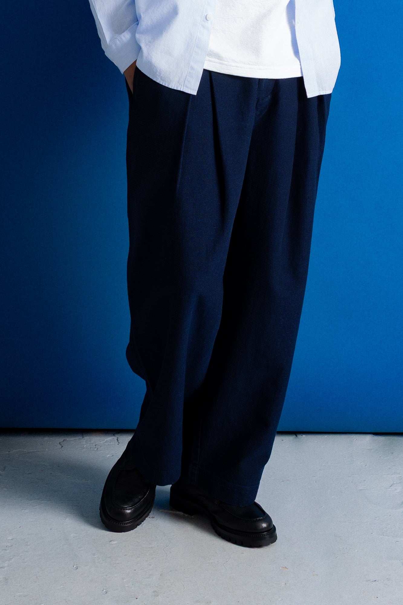 A person wearing loose dark navy pleated trousers, a white shirt, and black shoes, standing against a solid blue wall.