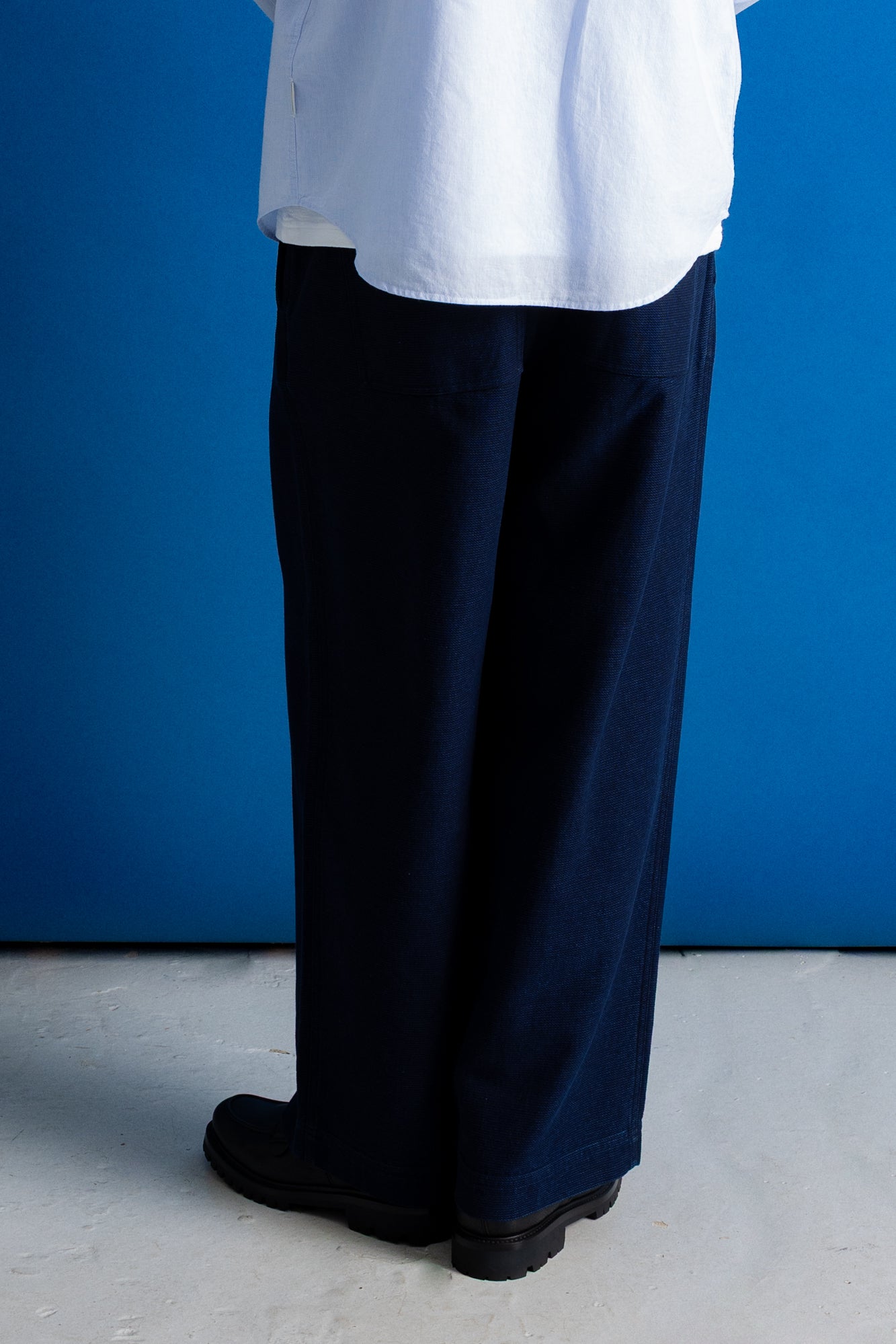 A person shown from the waist down wearing dark navy wide-leg trousers, a white shirt, and black boots, standing against a solid blue wall.