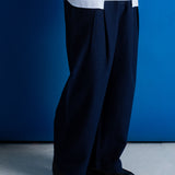 A person wearing loose dark navy pleated trousers, a white button-up shirt, and black shoes, standing against a solid blue wall and light gray floor.