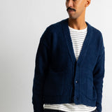 A man wearing a navy blue textured knit cardigan over a white and navy striped t-shirt, paired with an orange baseball cap and dark pants, standing against a plain off-white wall.
