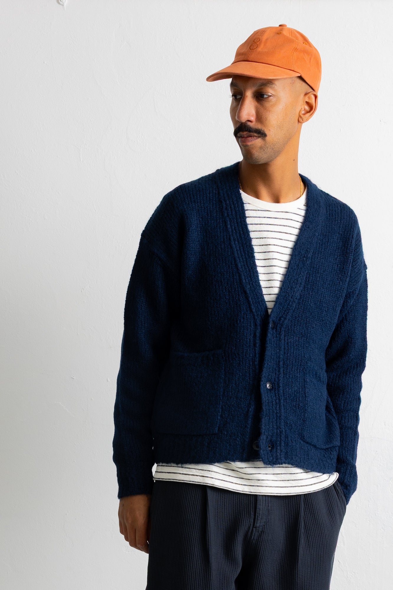 A man wearing a navy blue textured knit cardigan over a white and navy striped t-shirt, paired with an orange baseball cap and dark pants, standing against a plain off-white wall.