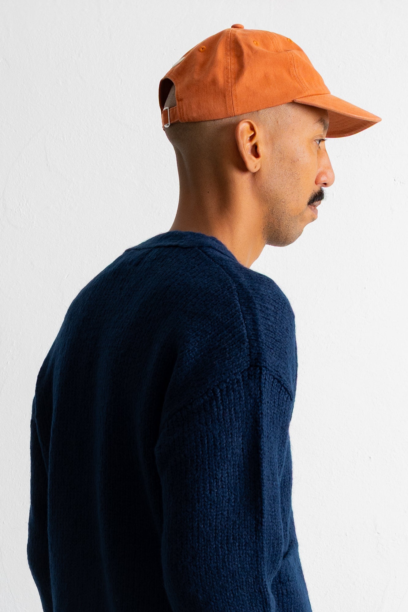 A man in profile wearing a dark navy knit sweater and an orange baseball cap, photographed against a plain white wall.