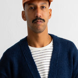 A man wearing an orange baseball cap, a navy blue knit cardigan, and a white t-shirt with thin black stripes, photographed against a plain white wall.