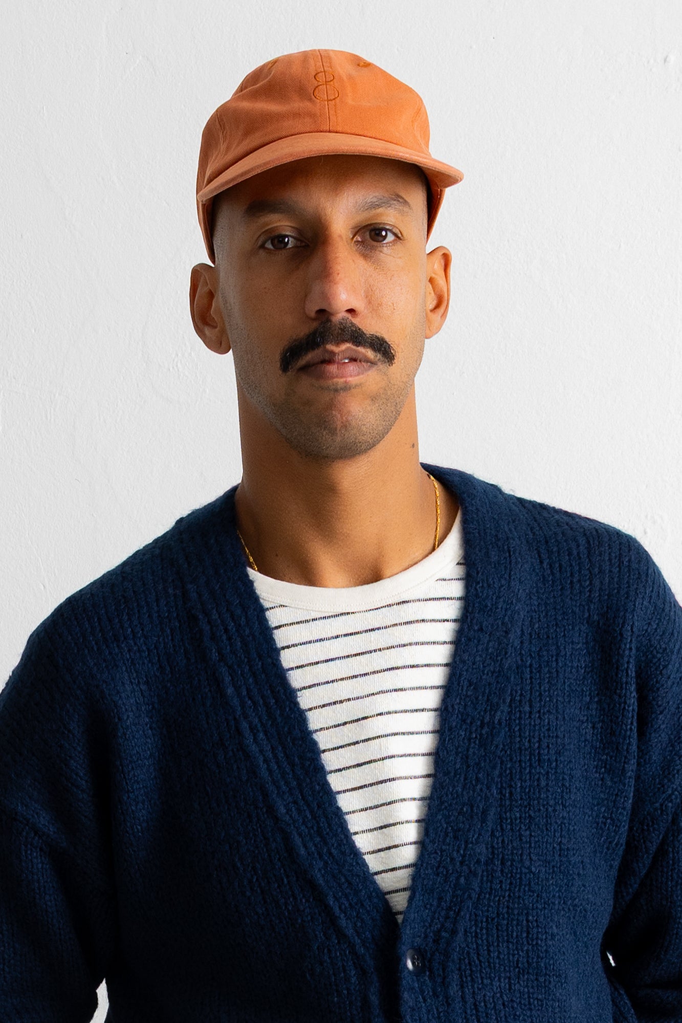 A man wearing an orange baseball cap, a navy blue knit cardigan, and a white t-shirt with thin black stripes, photographed against a plain white wall.