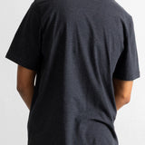 A man viewed from behind wearing a dark navy blue t-shirt with thin horizontal light stripes and teal pants, against a plain white wall.