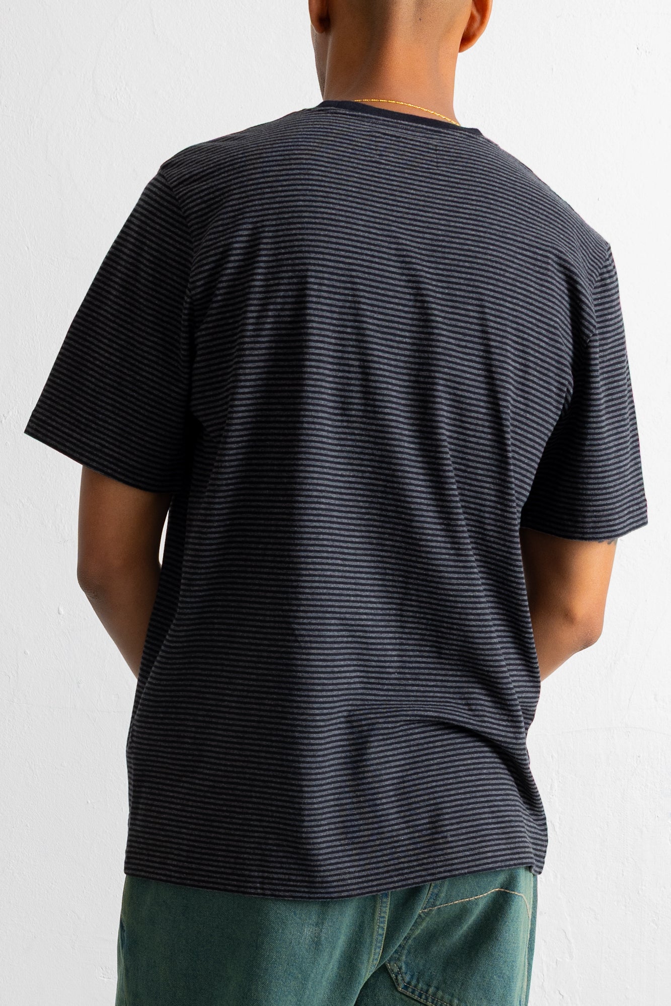 A man viewed from behind wearing a dark navy blue t-shirt with thin horizontal light stripes and teal pants, against a plain white wall.