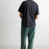 A man viewed from behind wearing a dark charcoal gray textured t-shirt, green pants, and black canvas sneakers, standing against a white wall.