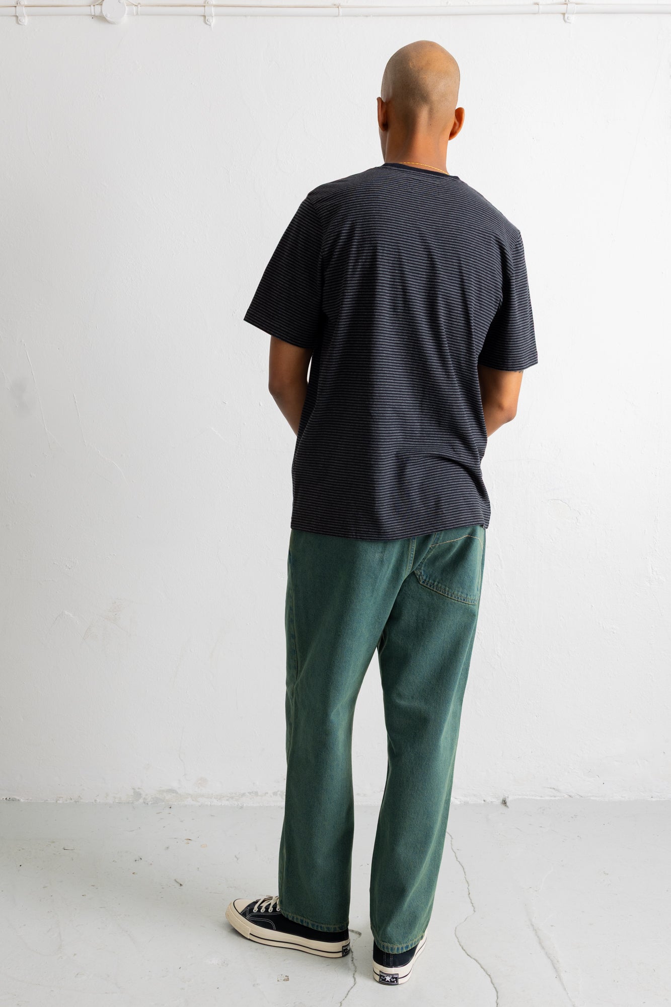 A man viewed from behind wearing a dark charcoal gray textured t-shirt, green pants, and black canvas sneakers, standing against a white wall.