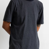 A man wearing a short-sleeve crewneck t-shirt with thin horizontal navy and black stripes, paired with teal jeans, shown from the waist up against a white wall.