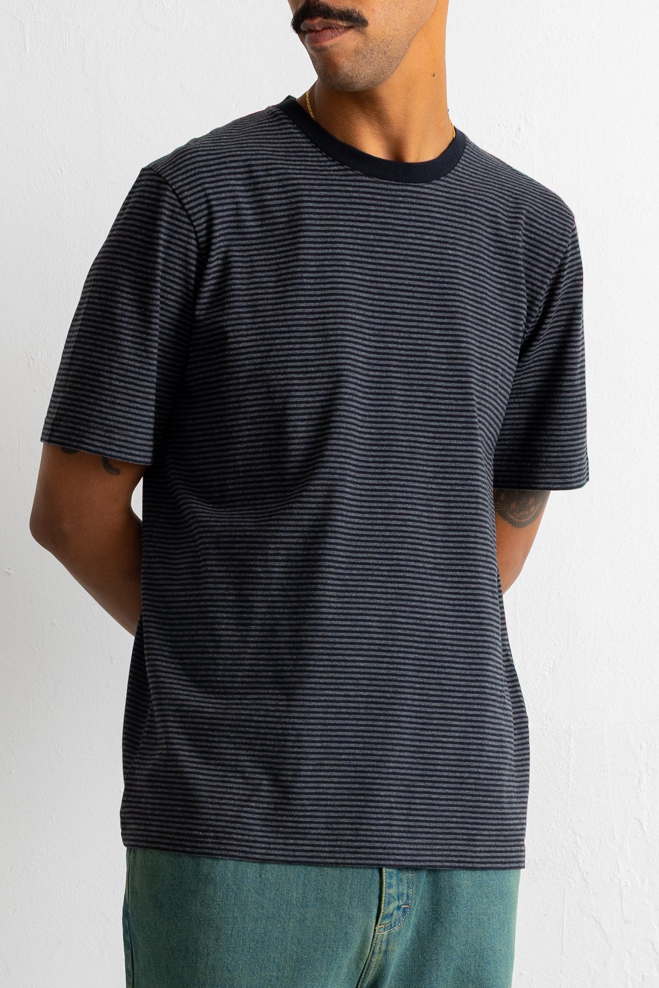A man wearing a short-sleeve crewneck t-shirt with thin horizontal navy and black stripes, paired with teal jeans, shown from the waist up against a white wall.