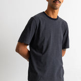 A man wearing a dark grey and black horizontally striped t-shirt with a black crew neck, paired with teal pants, standing against a plain light grey wall.