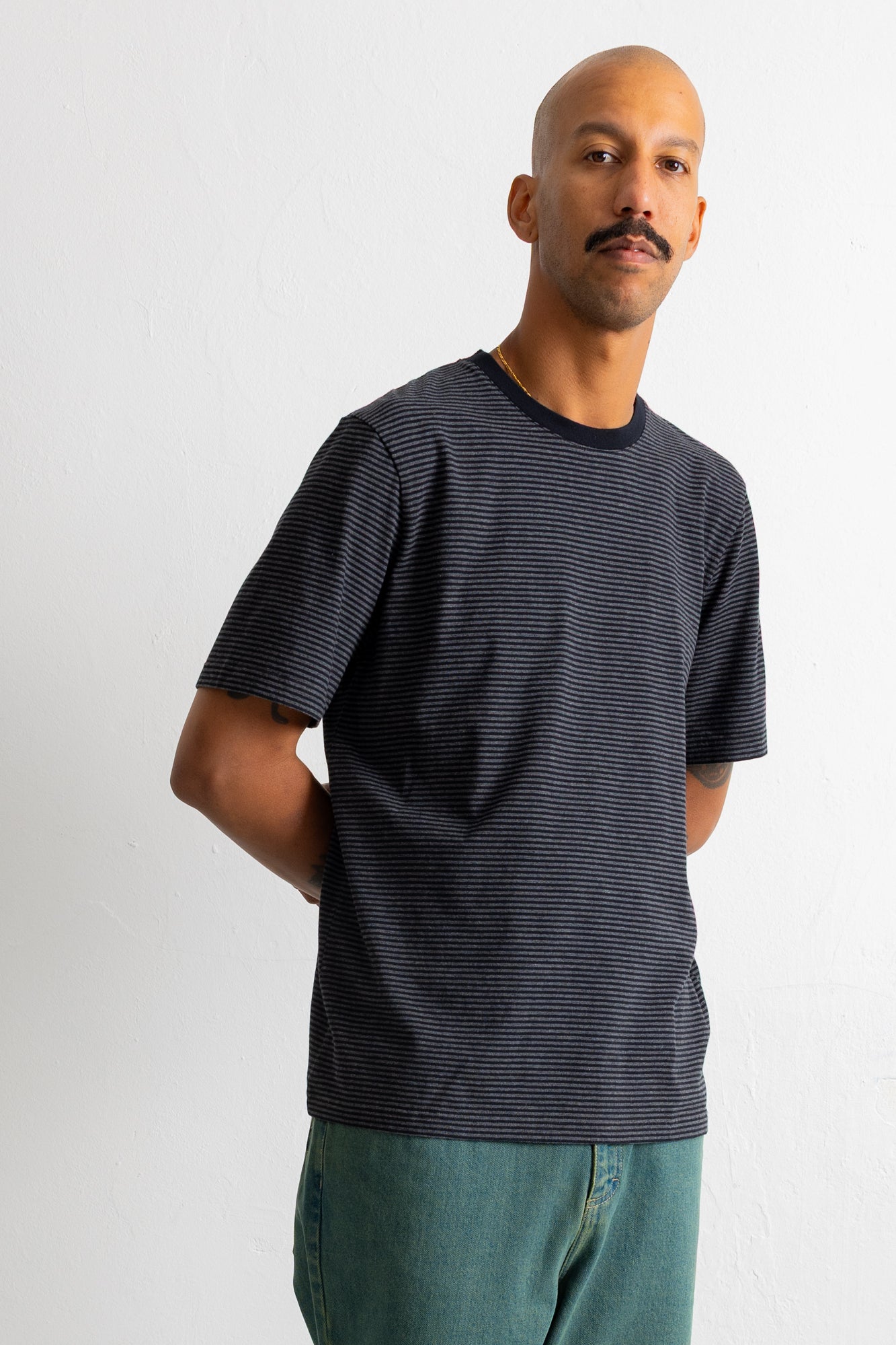 A man wearing a dark grey and black horizontally striped t-shirt with a black crew neck, paired with teal pants, standing against a plain light grey wall.