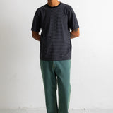 A man wearing a dark charcoal gray short-sleeve t-shirt, green pants, and black-and-white sneakers, standing against a plain white wall.