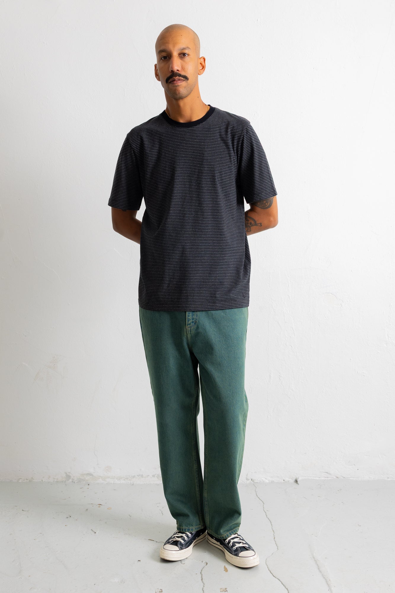 A man wearing a dark charcoal gray short-sleeve t-shirt, green pants, and black-and-white sneakers, standing against a plain white wall.