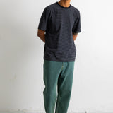 A man wearing a dark charcoal gray striped crewneck t-shirt, green pants, and black canvas sneakers, standing against a white wall.
