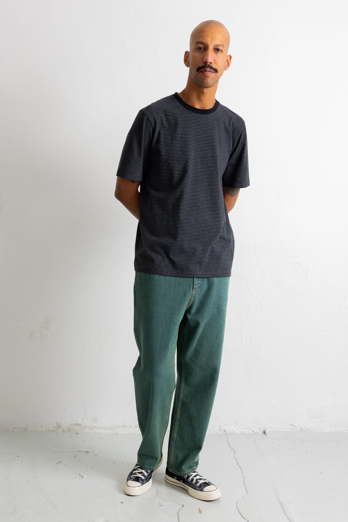 A man wearing a dark charcoal gray striped crewneck t-shirt, green pants, and black canvas sneakers, standing against a white wall.