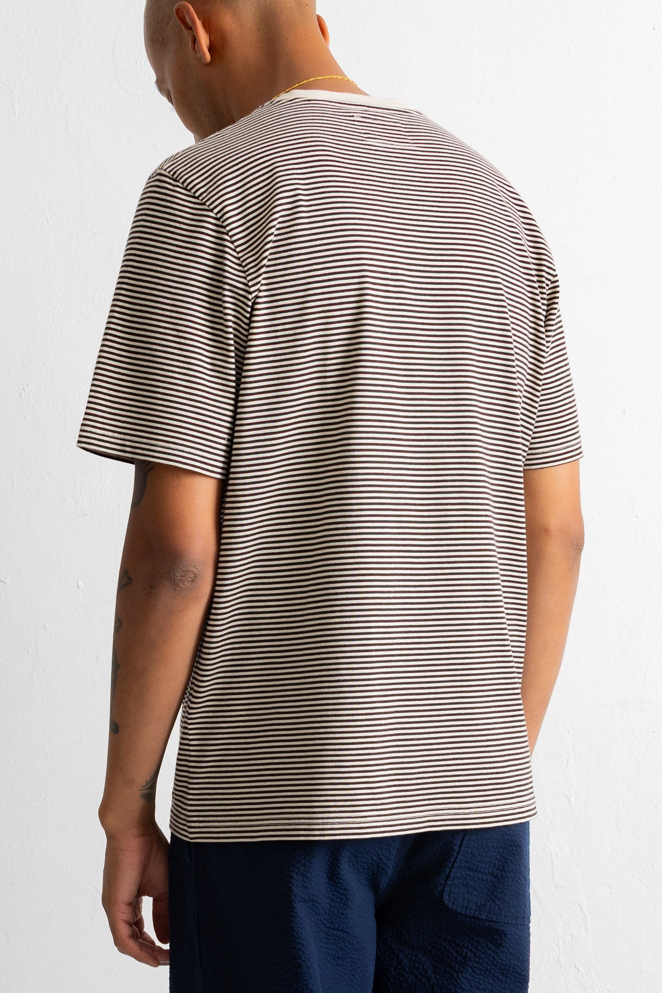 A man wearing a short-sleeve crewneck t-shirt with thin horizontal brown and beige stripes, shown from the back against a plain white wall.