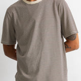 A man wearing a short-sleeve crewneck t-shirt with thin horizontal beige and black stripes, paired with dark blue pants, against a plain white background.