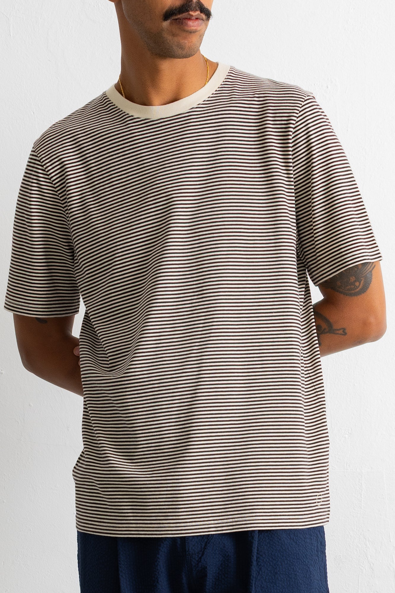A man wearing a short-sleeve crewneck t-shirt with thin horizontal beige and black stripes, paired with dark blue pants, against a plain white background.