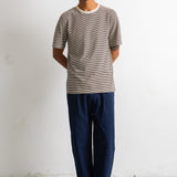 A man wearing a short-sleeve crewneck t-shirt with fine horizontal stripes in taupe and gray, paired with dark navy trousers and dark brown leather shoes, standing against a white studio wall.