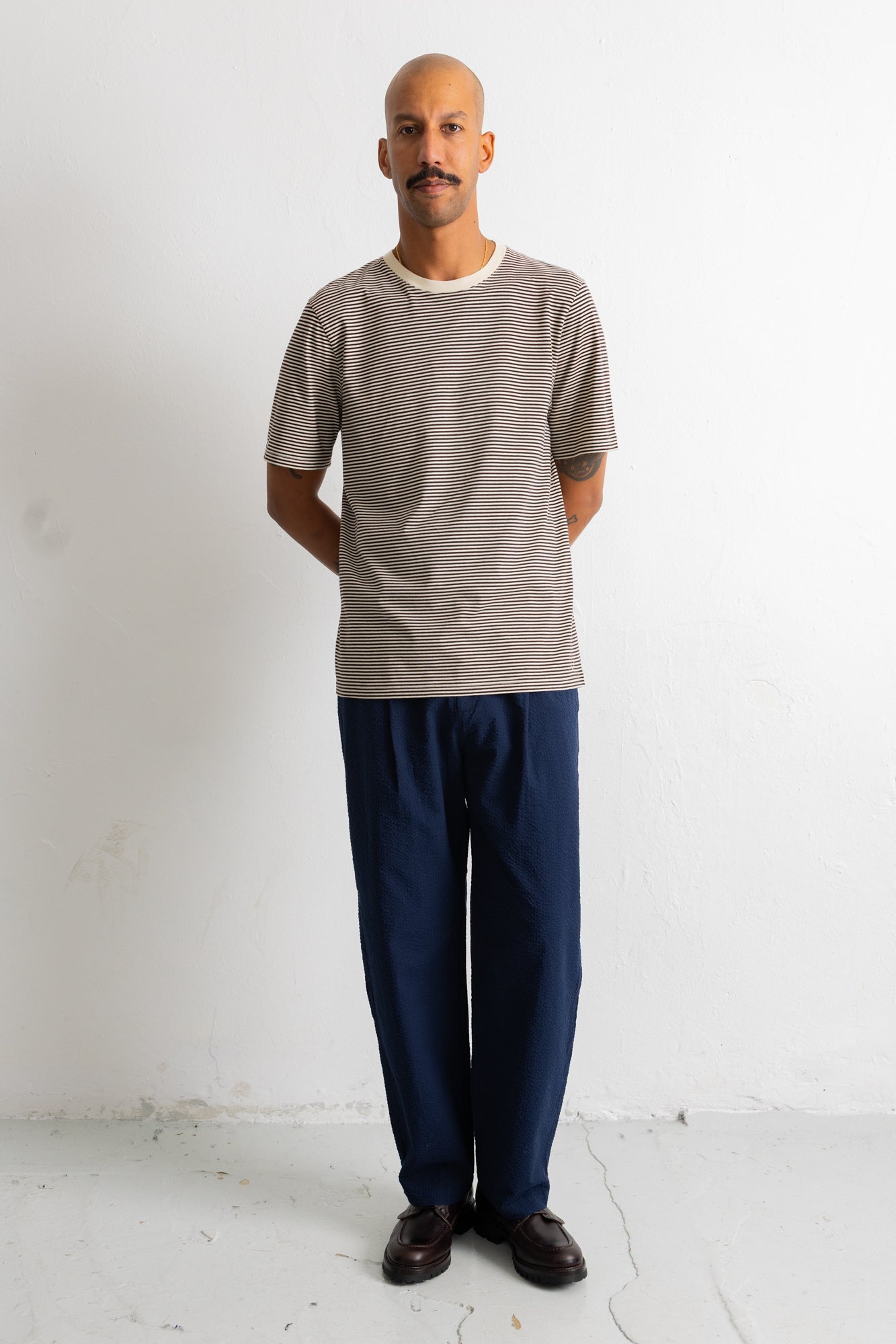 A man wearing a short-sleeve crewneck t-shirt with fine horizontal stripes in taupe and gray, paired with dark navy trousers and dark brown leather shoes, standing against a white studio wall.