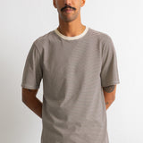 A man wearing a short-sleeve crewneck t-shirt with thin horizontal beige and dark gray stripes, paired with navy blue pants, standing against a plain white wall.