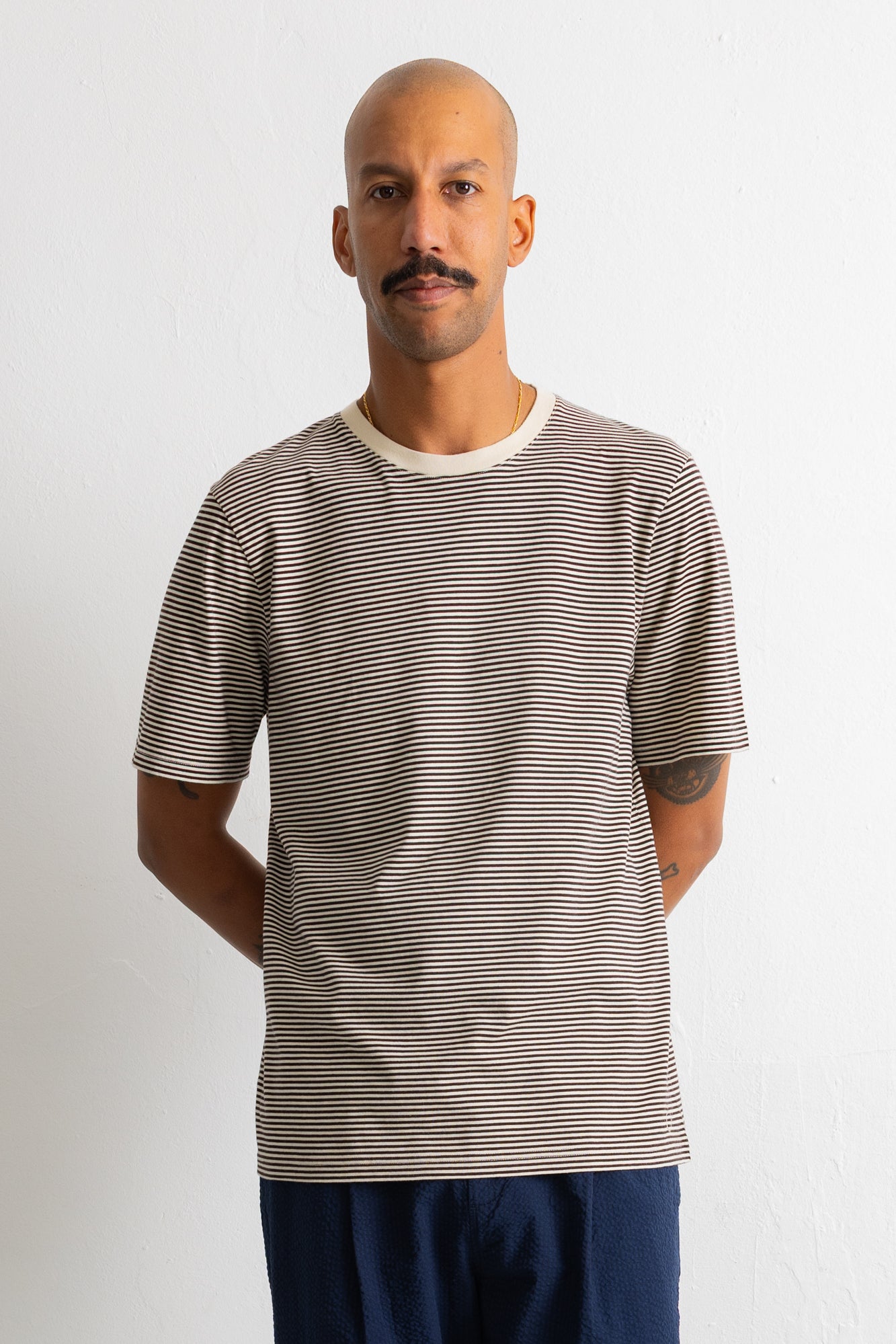 A man wearing a short-sleeve crewneck t-shirt with thin horizontal beige and dark gray stripes, paired with navy blue pants, standing against a plain white wall.
