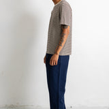 A man stands in profile wearing a short-sleeve striped t-shirt in gray and white, paired with dark blue trousers and dark brown shoes, against a plain white studio wall.