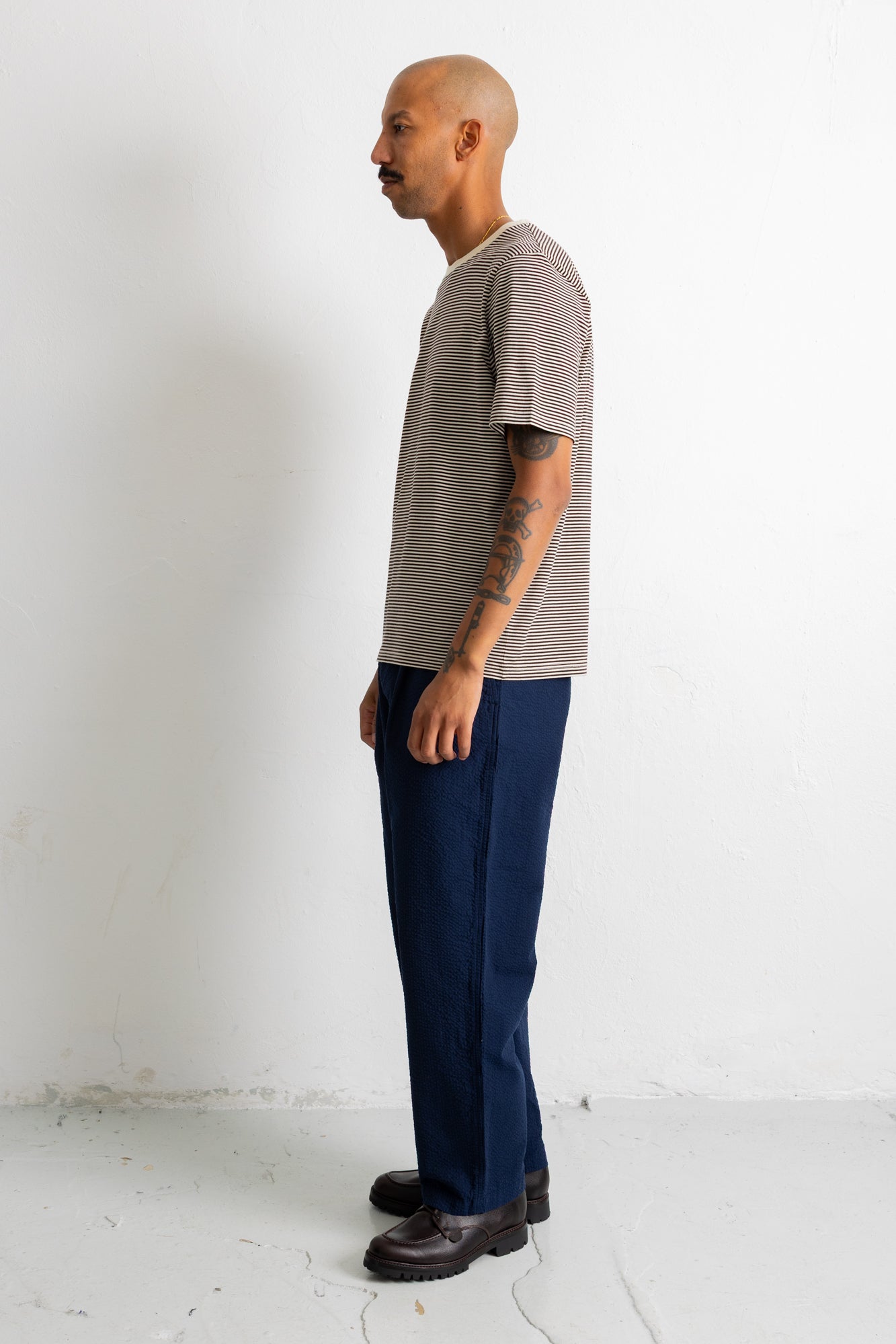 A man stands in profile wearing a short-sleeve striped t-shirt in gray and white, paired with dark blue trousers and dark brown shoes, against a plain white studio wall.