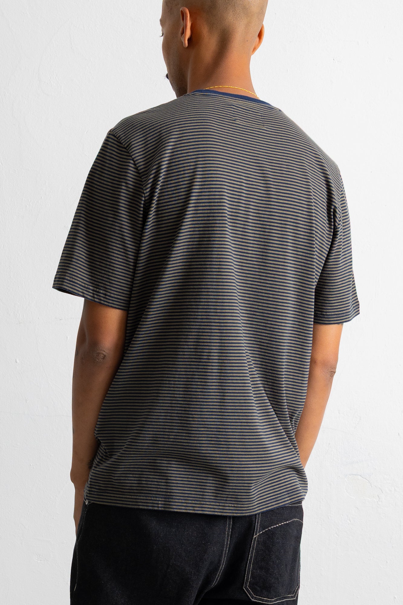 A man viewed from behind wearing a dark green and black horizontally striped t-shirt and dark jeans against a plain white background.