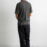 A man viewed from behind wearing a dark green short-sleeve striped t-shirt, black jeans, and black canvas sneakers, standing against a plain white wall.