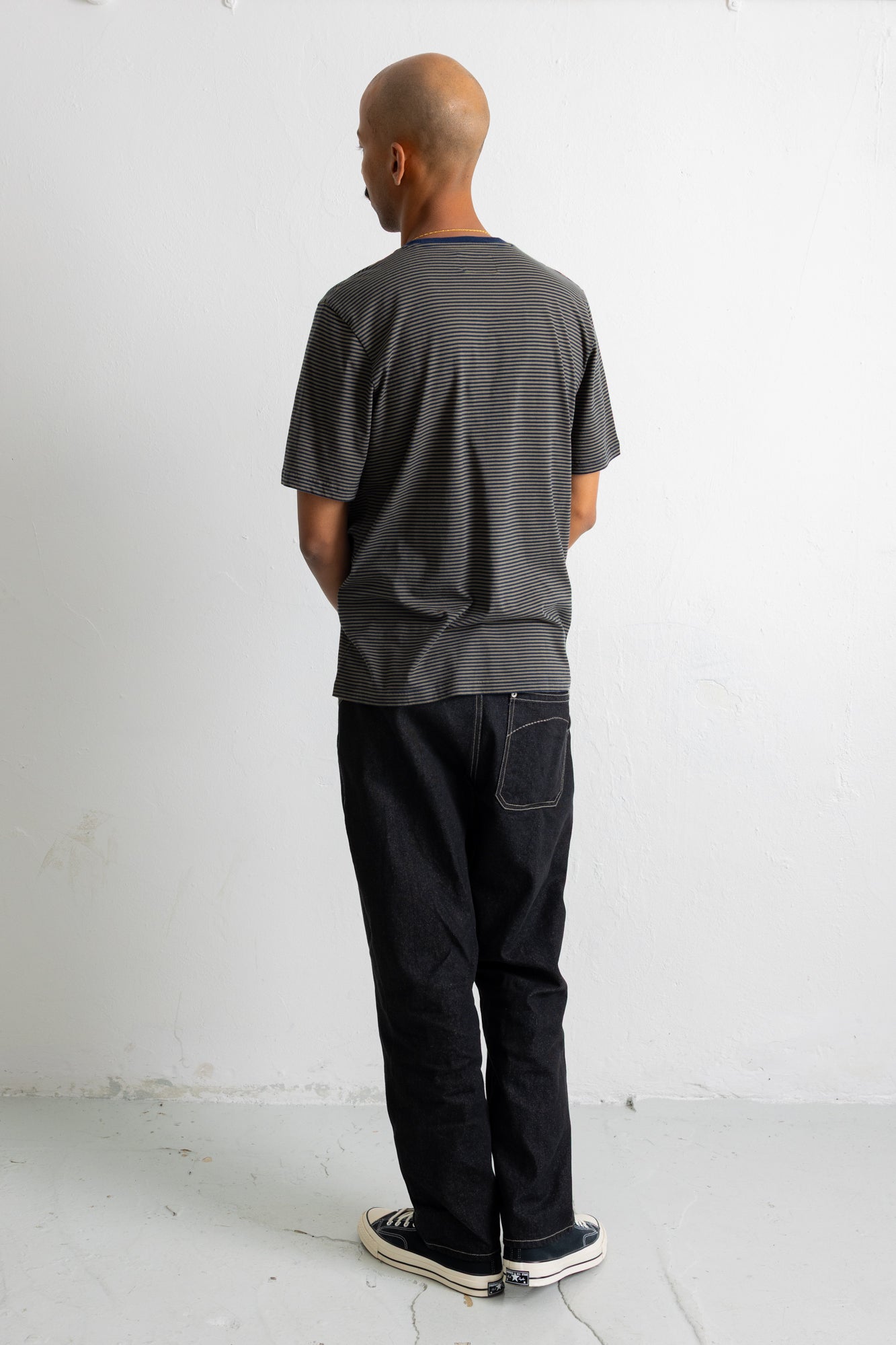 A man viewed from behind wearing a dark green short-sleeve striped t-shirt, black jeans, and black canvas sneakers, standing against a plain white wall.