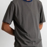 A man wearing a dark green and black horizontally striped t-shirt with a contrasting navy blue crew neck, shown from the waist up against a plain light gray background