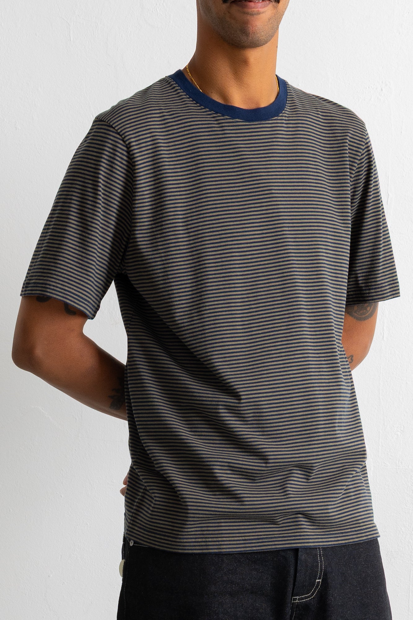 A man wearing a dark green and black horizontally striped t-shirt with a contrasting navy blue crew neck, shown from the waist up against a plain light gray background