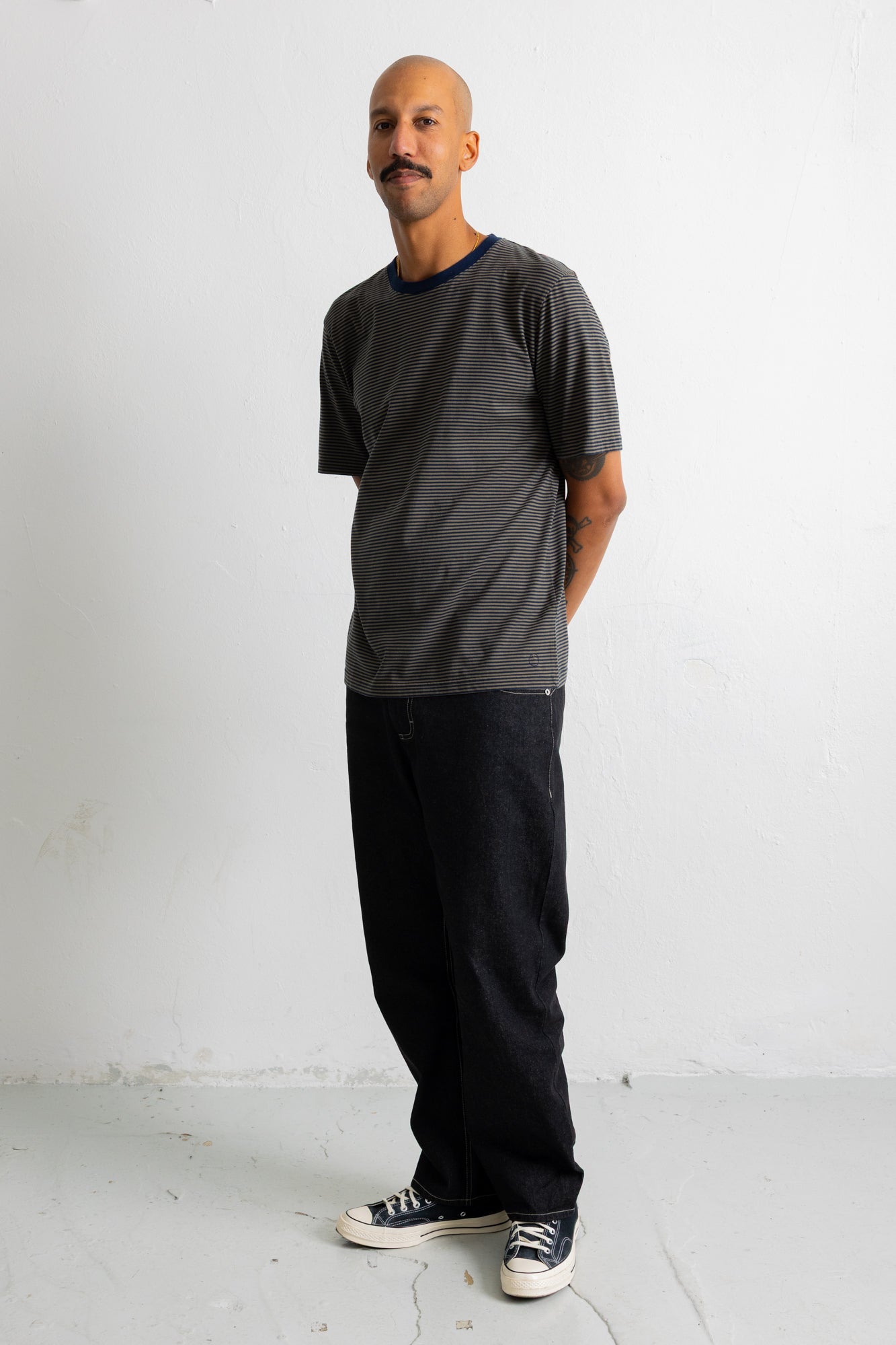A man wearing a dark green short-sleeve t-shirt with a dark navy collar, black pants, and black canvas sneakers, standing against a white studio wall.