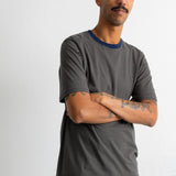 A man wearing a dark green and black horizontally striped t-shirt with a navy blue crew neck, standing with arms crossed against a plain light gray background.