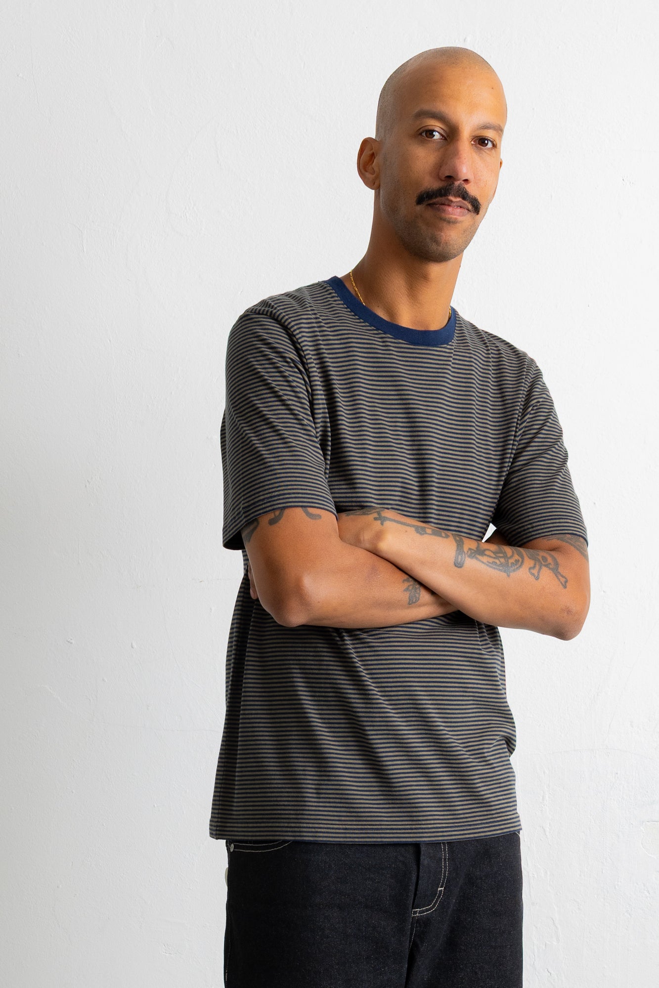 A man wearing a dark green and black horizontally striped t-shirt with a navy blue crew neck, standing with arms crossed against a plain light gray background.