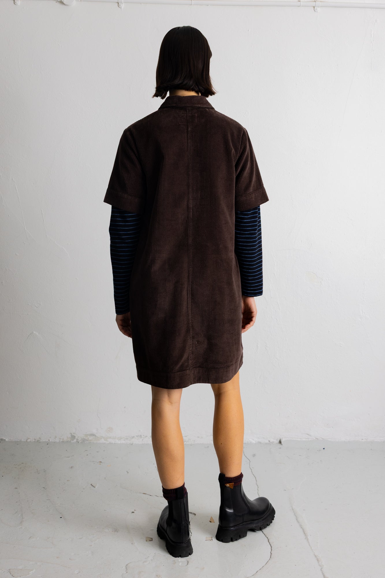 A model wearing a dark brown corduroy short-sleeve collared pullover over a blue and black striped long-sleeve shirt, set against a plain light gray background.