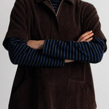 A model wearing a dark brown corduroy short-sleeve collared pullover over a blue and black striped long-sleeve shirt, set against a plain light gray background.