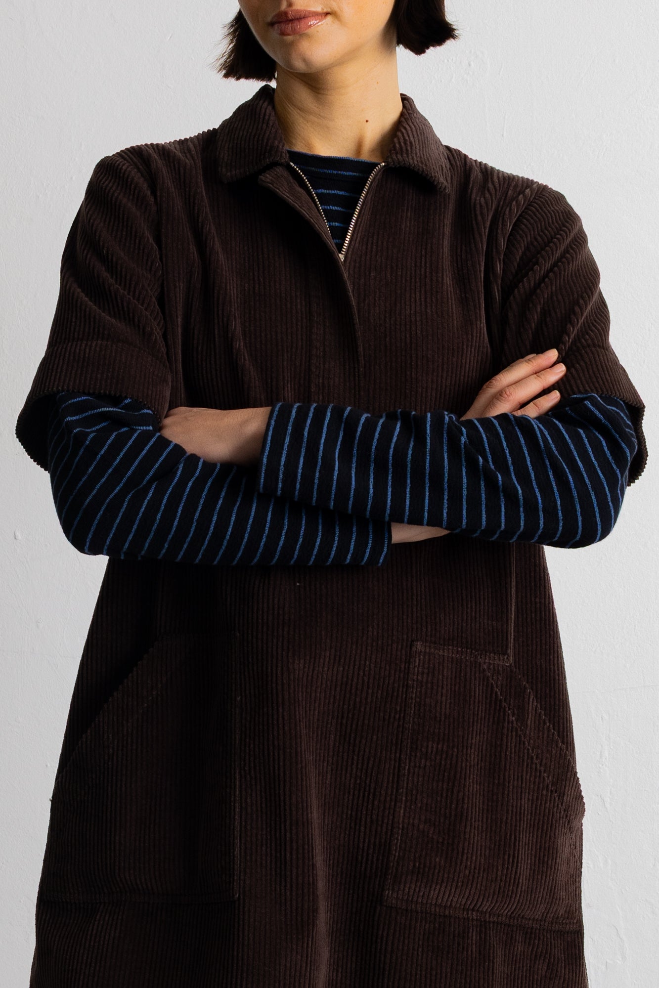 A model wearing a dark brown corduroy short-sleeve collared pullover over a blue and black striped long-sleeve shirt, set against a plain light gray background.