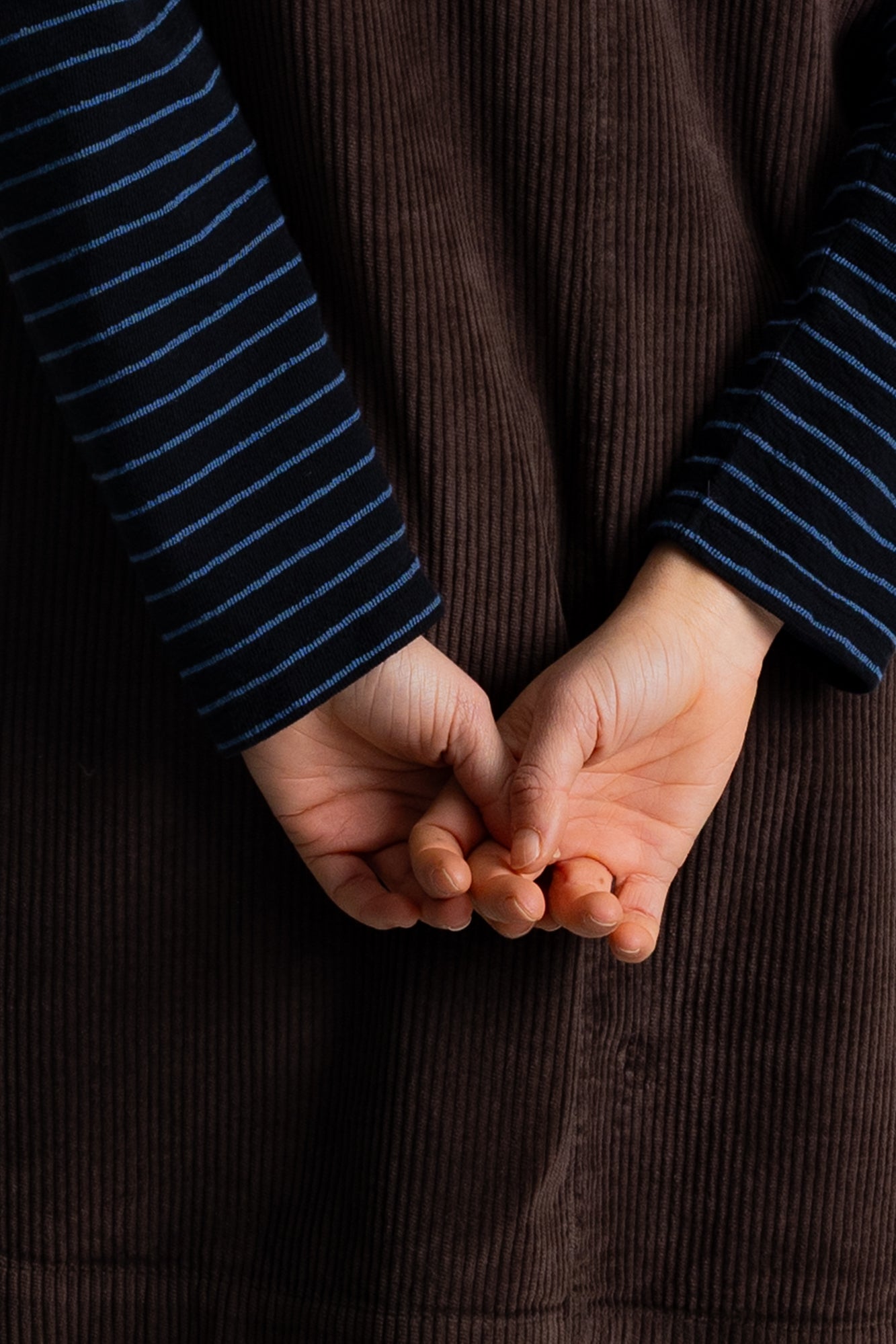 Close-up of a person's hands clasped behind a dark brown corduroy garment, with navy and blue striped sleeves visible.