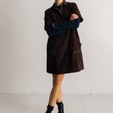 A model stands in a studio wearing a dark brown corduroy mini coat over a navy and black striped long-sleeve top, paired with black ankle boots.