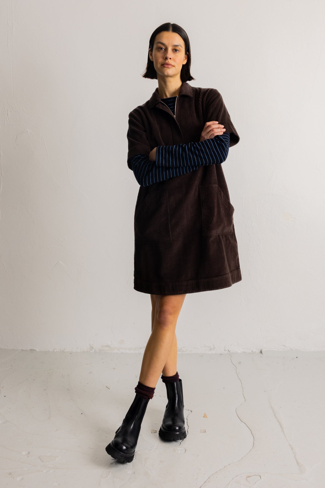 A model stands in a studio wearing a dark brown corduroy mini coat over a navy and black striped long-sleeve top, paired with black ankle boots.